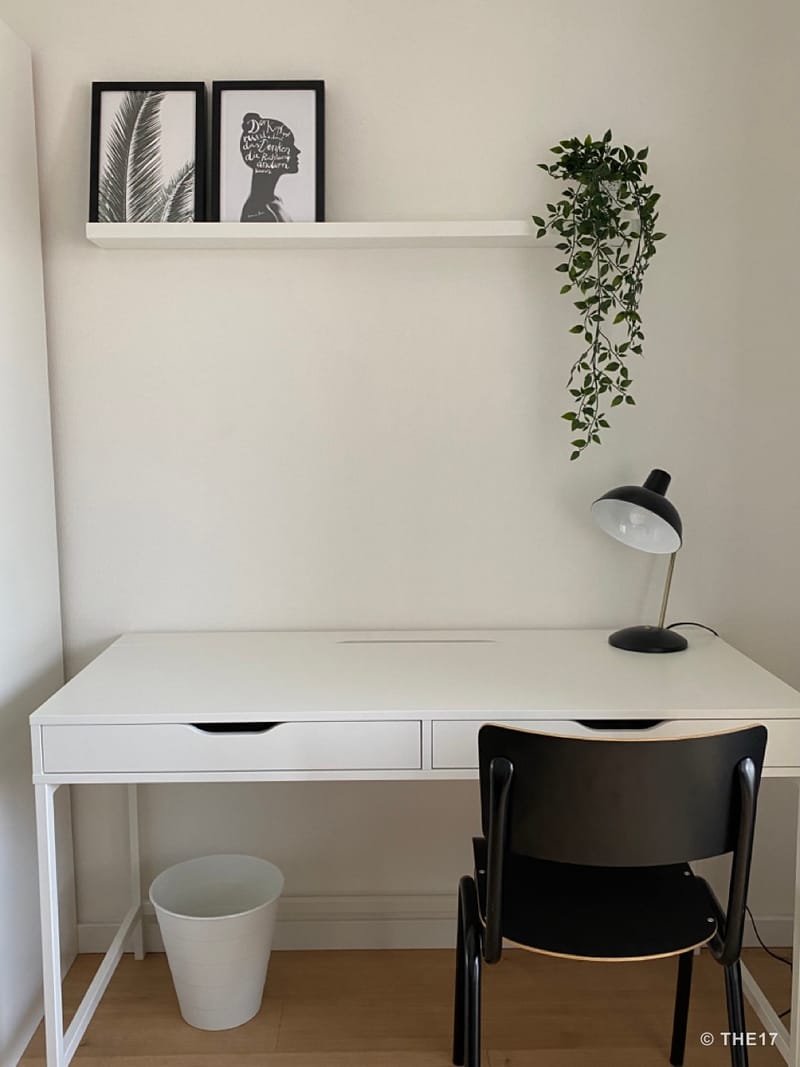 Weißer Schreibtisch mit zwei Schubladen, schwarzer Stuhl, schwarze Lampe, weiße Wand mit zwei gerahmten Bildern und hängender Pflanze.