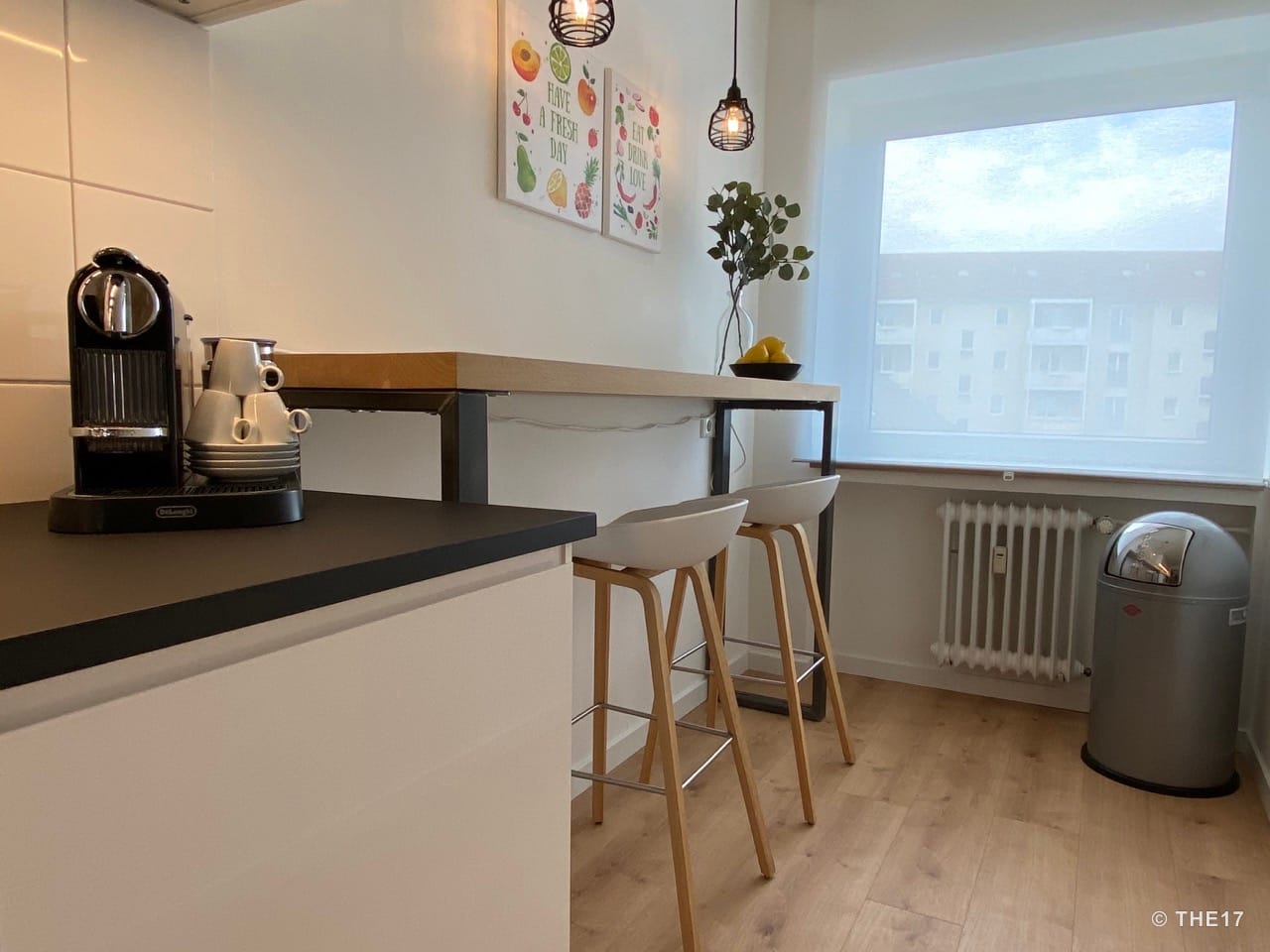 Kleine Küche mit Holztheke, zwei weißen Barhockern, Kaffeemaschine auf schwarzer Arbeitsplatte und Fenster mit weißem Rollo.