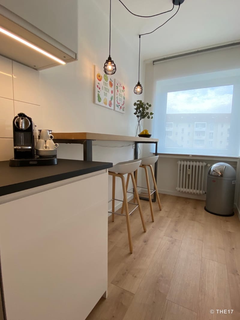 Moderne Küche mit schwarzer Arbeitsplatte, Holzbar mit zwei Hockern, zwei Hängeleuchten und großem Fenster mit Jalousie.