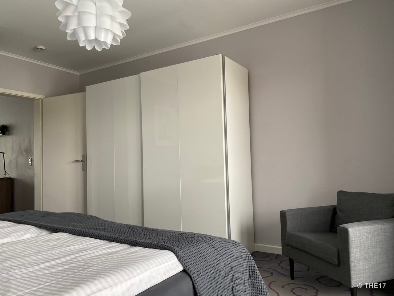 Schlafzimmer mit weißem Schrank, grauem Sessel, weißer Tür und modernem Deckenlicht.