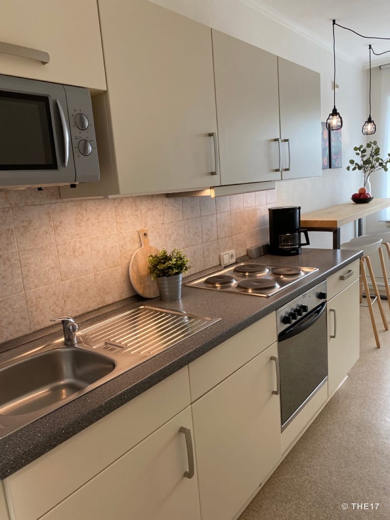 Moderne Küche mit Einbauherd, Spüle, Mikrowelle, Kaffeemaschine und Bar mit Hockern, helle Schränke und Pendelleuchten.