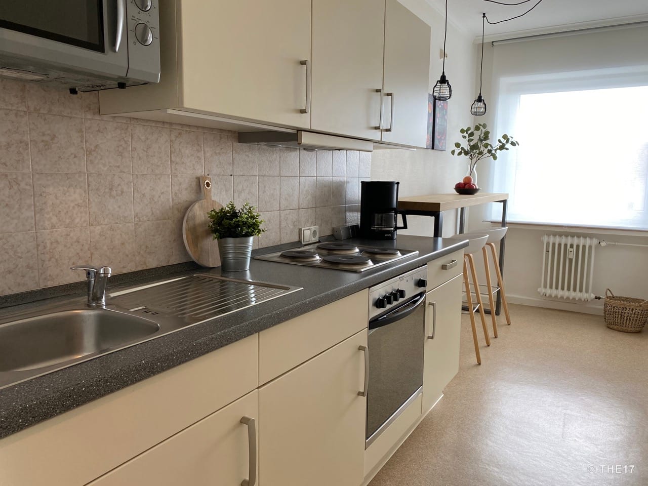 Moderne Küche mit Spüle, Herd, Kaffeemaschine und hoher Theke mit Barhockern neben hellem Fenster.