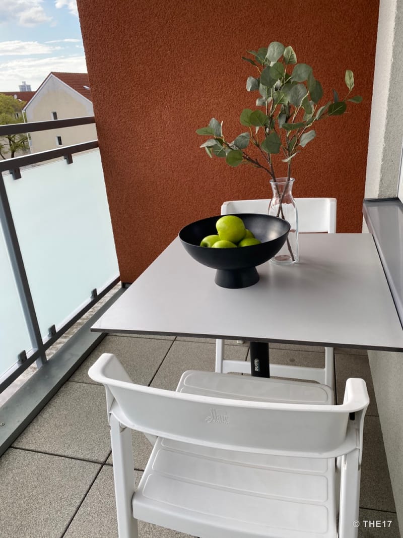 Balkon mit weißem Tisch und Stühlen, schwarzer Schale mit grünen Äpfeln und Glasvase mit grünen Zweigen.