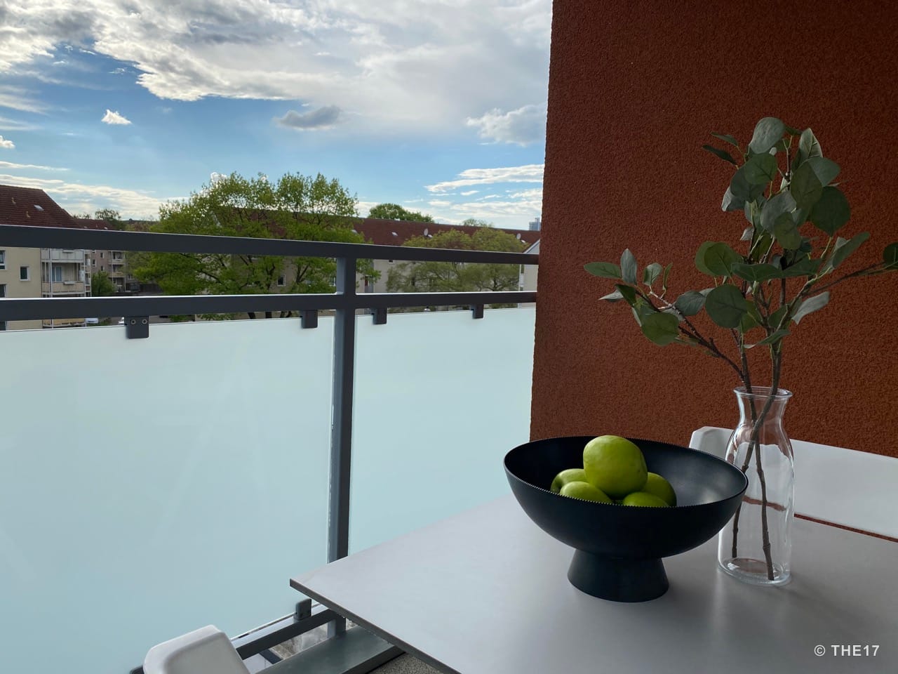 Balkon mit mattierter Glaspaneele, Tisch mit schwarzer Schale gefüllt mit grünen Äpfeln und Glasvase mit Zweigen vor roter Wand.