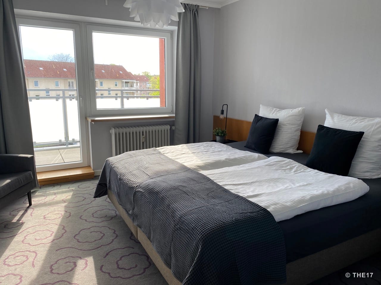Schlafzimmer mit Doppelbett, weißen und schwarzen Kissen, schmalem Nachttisch mit Lampe und Pflanze, Zugang zum Balkon.