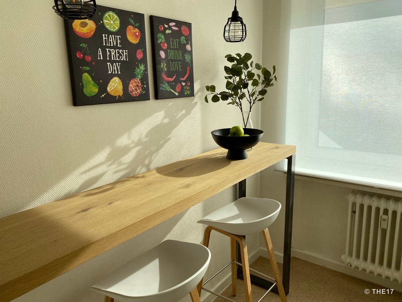 Heller Essbereich mit hohem Holztisch, zwei weißen Barhockern, schwarzer Obstschale und Wanddekoration mit Sprüchen.