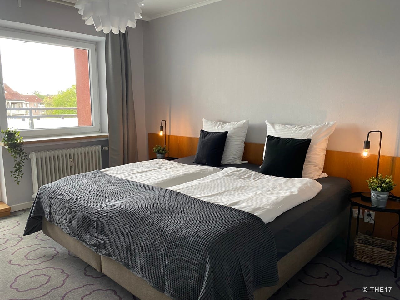 Schlafzimmer mit Doppelbett, weißen und schwarzen Kissen, grauer Bettdecke, zwei Nachttischen mit Lampen und Topfpflanzen, Fenster mit grauem Vorhang.