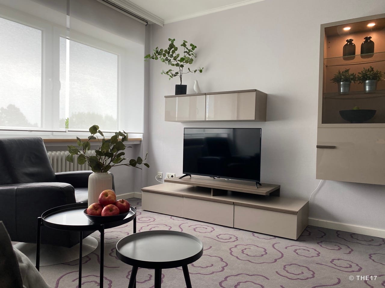 Moderner Wohnraum mit schwarzem Ledersessel, grauem Teppich mit lila Mustern, TV-Möbel in Beige und großen Fenstern mit weißen Jalousien.
