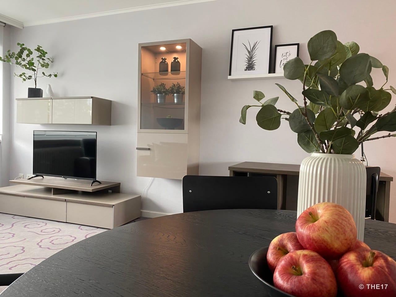 Esszimmer mit rundem schwarzem Tisch, Äpfeln in einer Schale, grüner Pflanze, TV auf Sideboard und hängendem Regal.