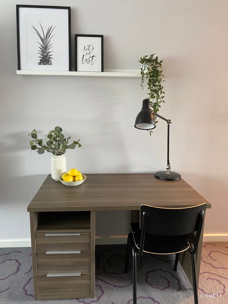 Pult mit drei Schubladen, schwarzem Stuhl, Vase mit grünen Blättern, Schale mit Zitronen und schwarzer Lampe vor weißer Wand.