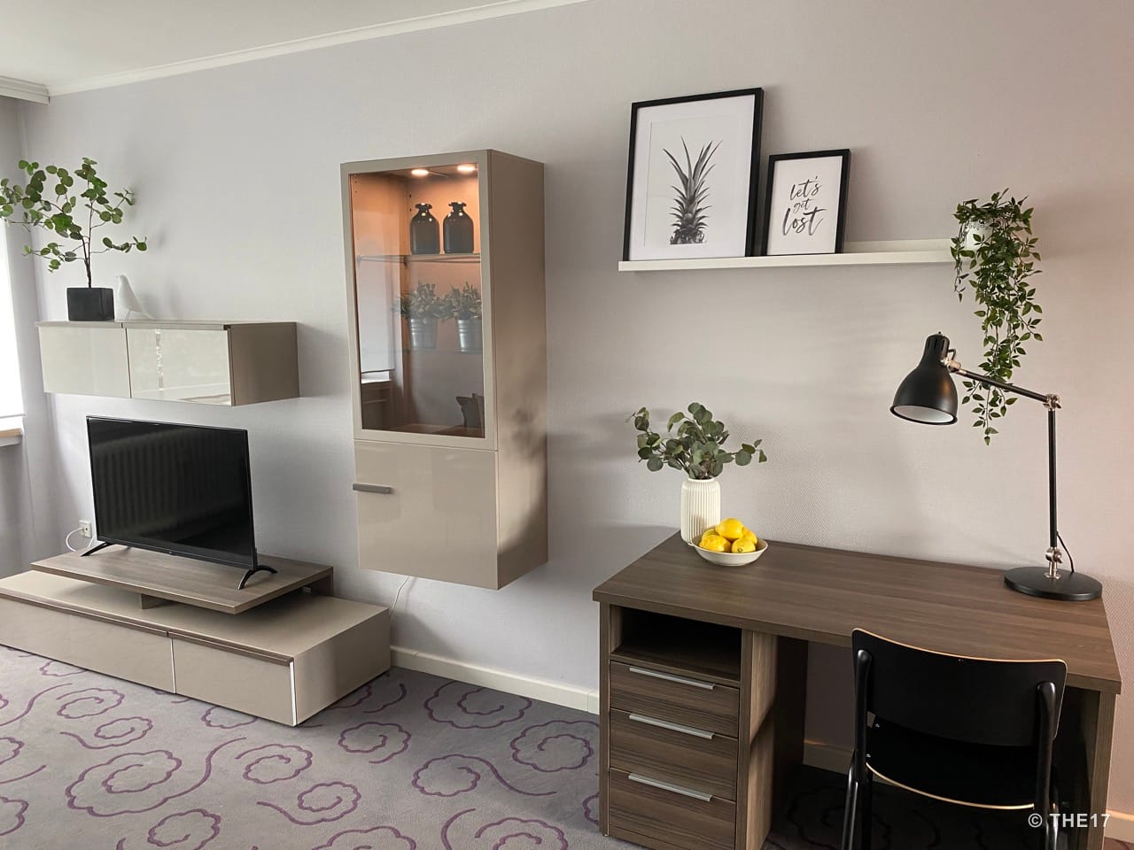 Moderner Wohnbereich mit Wandregalen, TV, Glasvitrine, Schreibtisch mit Lampe, Pflanze und Bildern an der Wand.