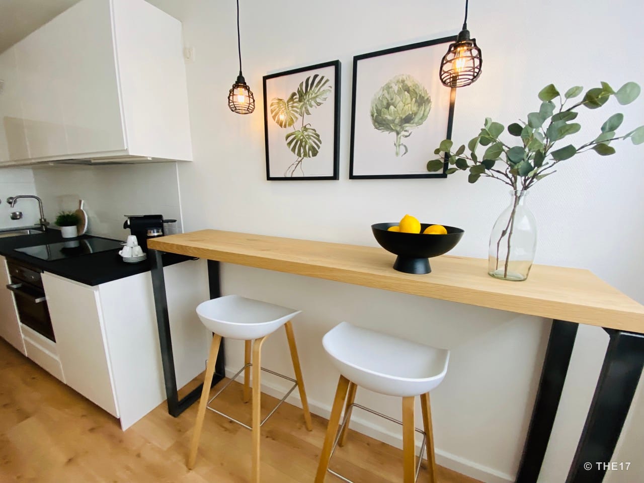Modern dining area with bar table, two white stools, two botanical prints, and vase with leafy branches.
