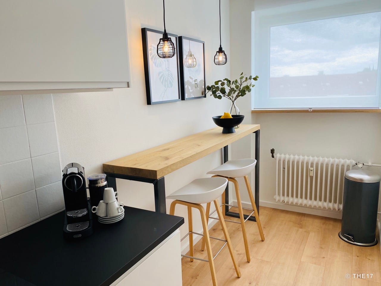 Small kitchen corner with black coffee machine, wooden bar table with two white high stools, hanging lights and window with blind.