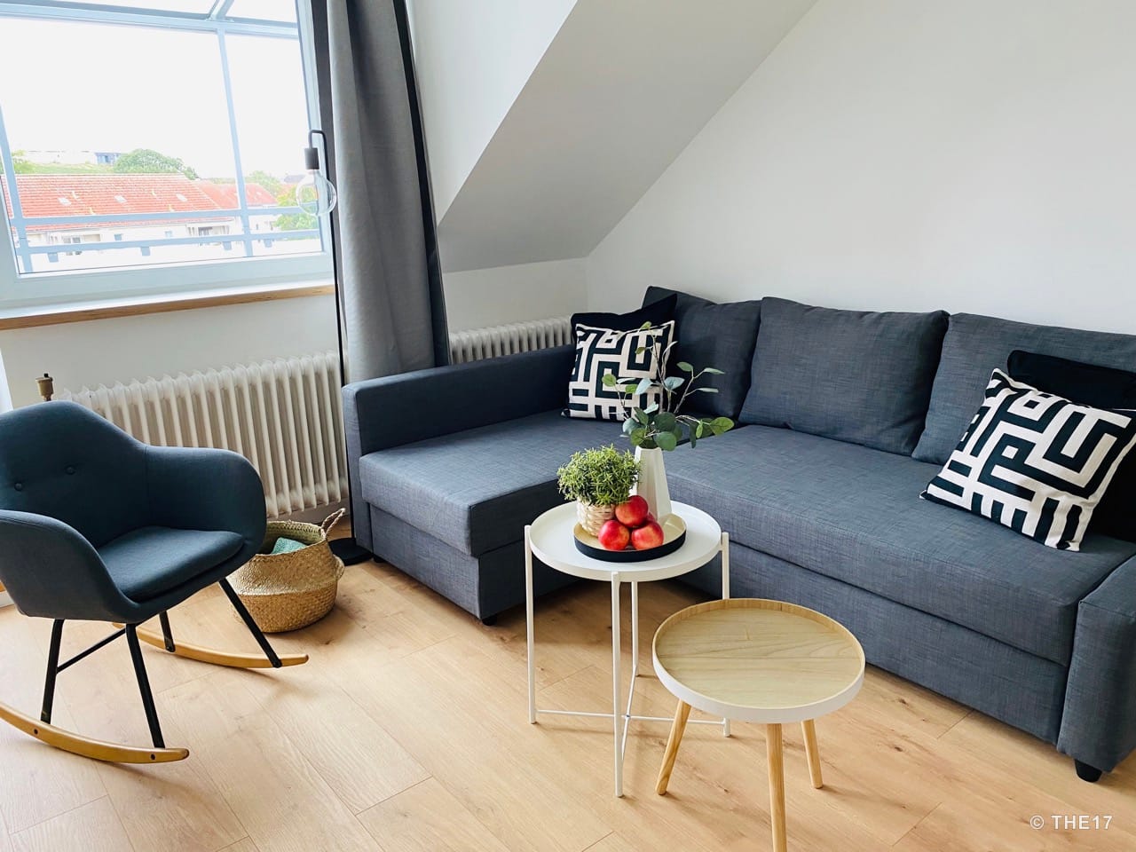 Modern living room with a gray sofa, black and white cushions, red apples on tables, and a rocking chair by the window.