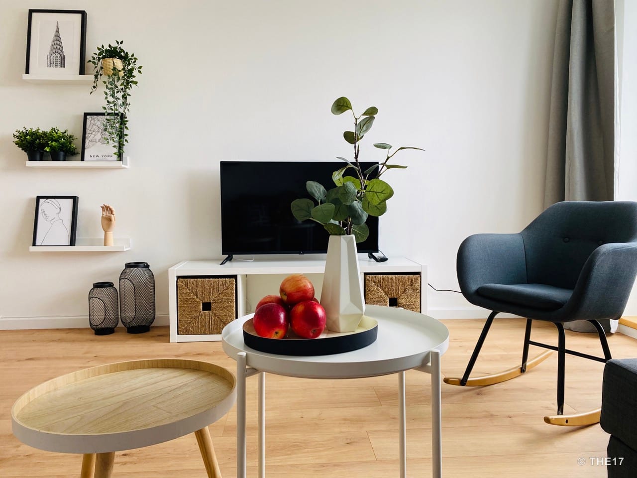 Hell eingerichtetes Wohnzimmer mit grauem Schaukelstuhl, zwei Couchtischen, TV auf weißem TV-Schrank und dekorativen Wandregalen.