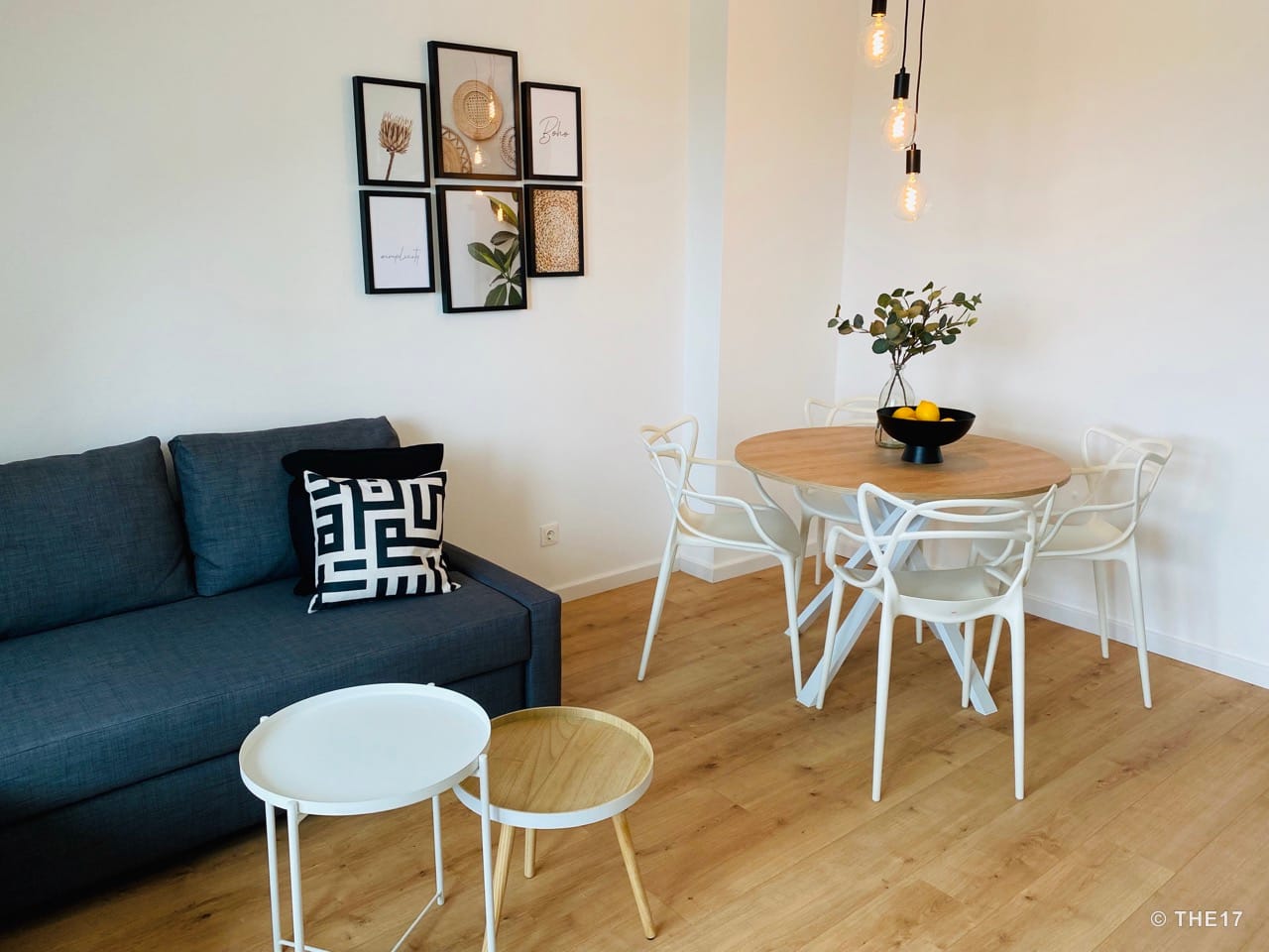 Modern living room with dark grey sofa, wooden dining table, white chairs, and minimalist wall art.