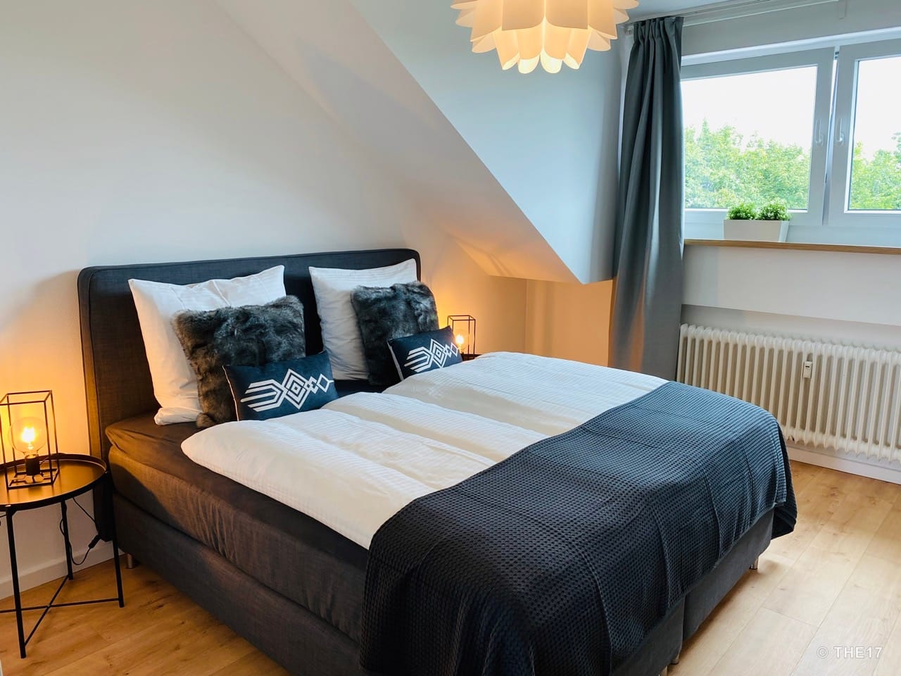 Bedroom with gray double bed, white pillows, small black cushions, two bedside lamps, and window with gray curtain.