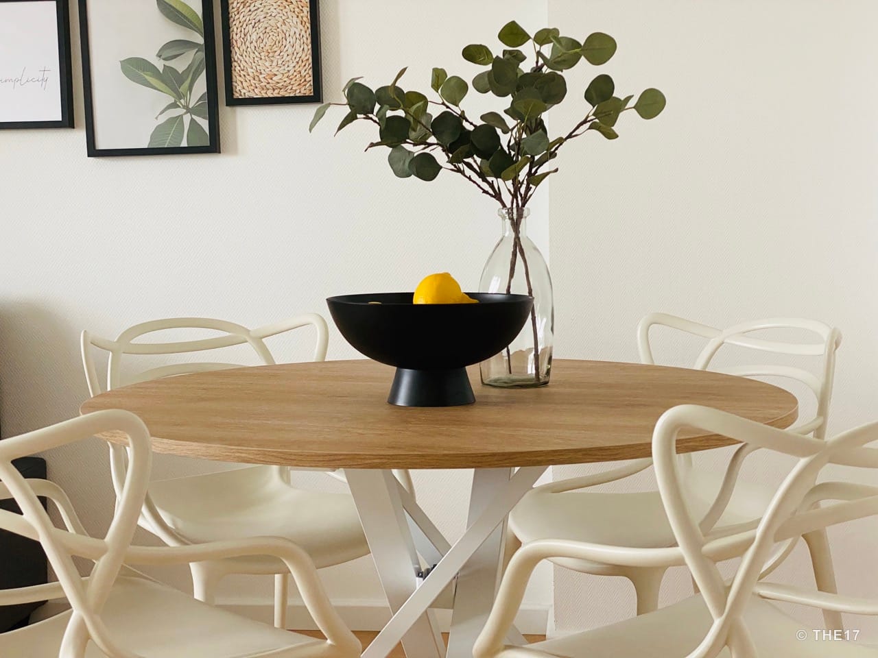 Modern round wooden table with white metal base, four white chairs, black fruit bowl with lemon in a bright room.