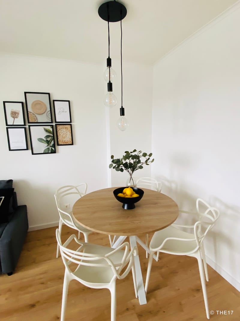 Modern dining area with round wooden table, white chairs, black bowl with lemons, framed wall art, and hanging light bulbs.