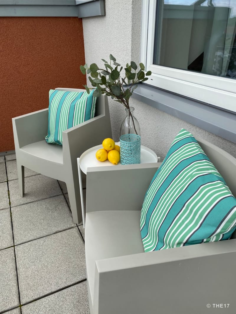 Two grey chairs with striped cushions on balcony, small white table with lemons and plant in glass bottle.