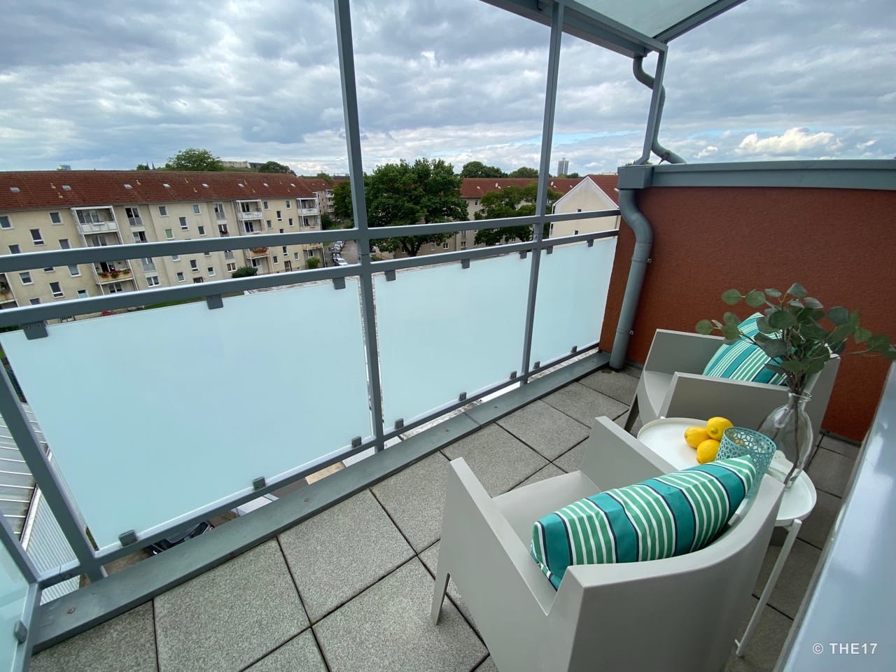 Balcony with two chairs, striped cushions, small white table with lemons and vase, view of neighboring buildings.