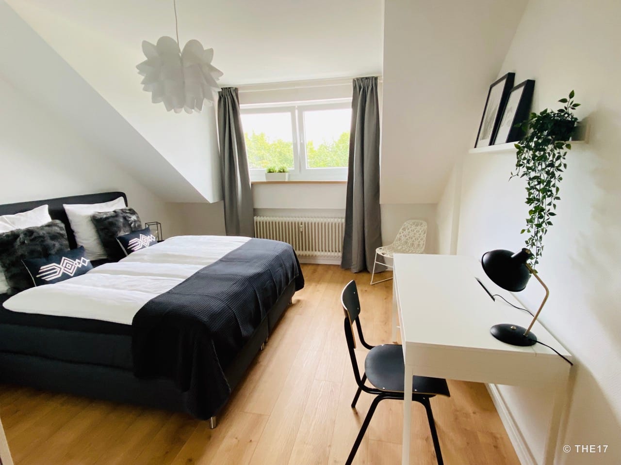 Bedroom with double bed, white desk with black chair, window with gray curtains, and wooden floor.