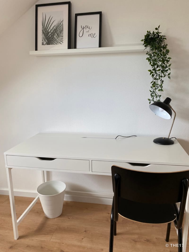 White desk with two drawers, black chair, black lamp, and hanging plant decoration on a white wall with shelves.