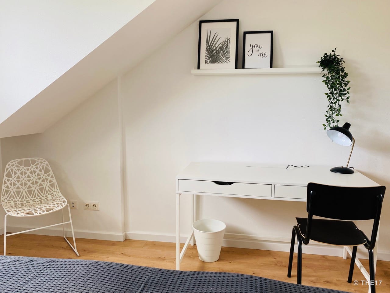 White desk with black chair, framed pictures, and green plant on white wall under slanted ceiling.