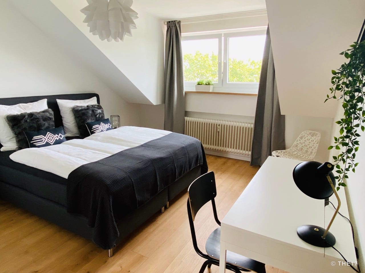 Bedroom with double bed, black pillows, white bedding, desk with chair, gray curtains, and window showing green trees outside.