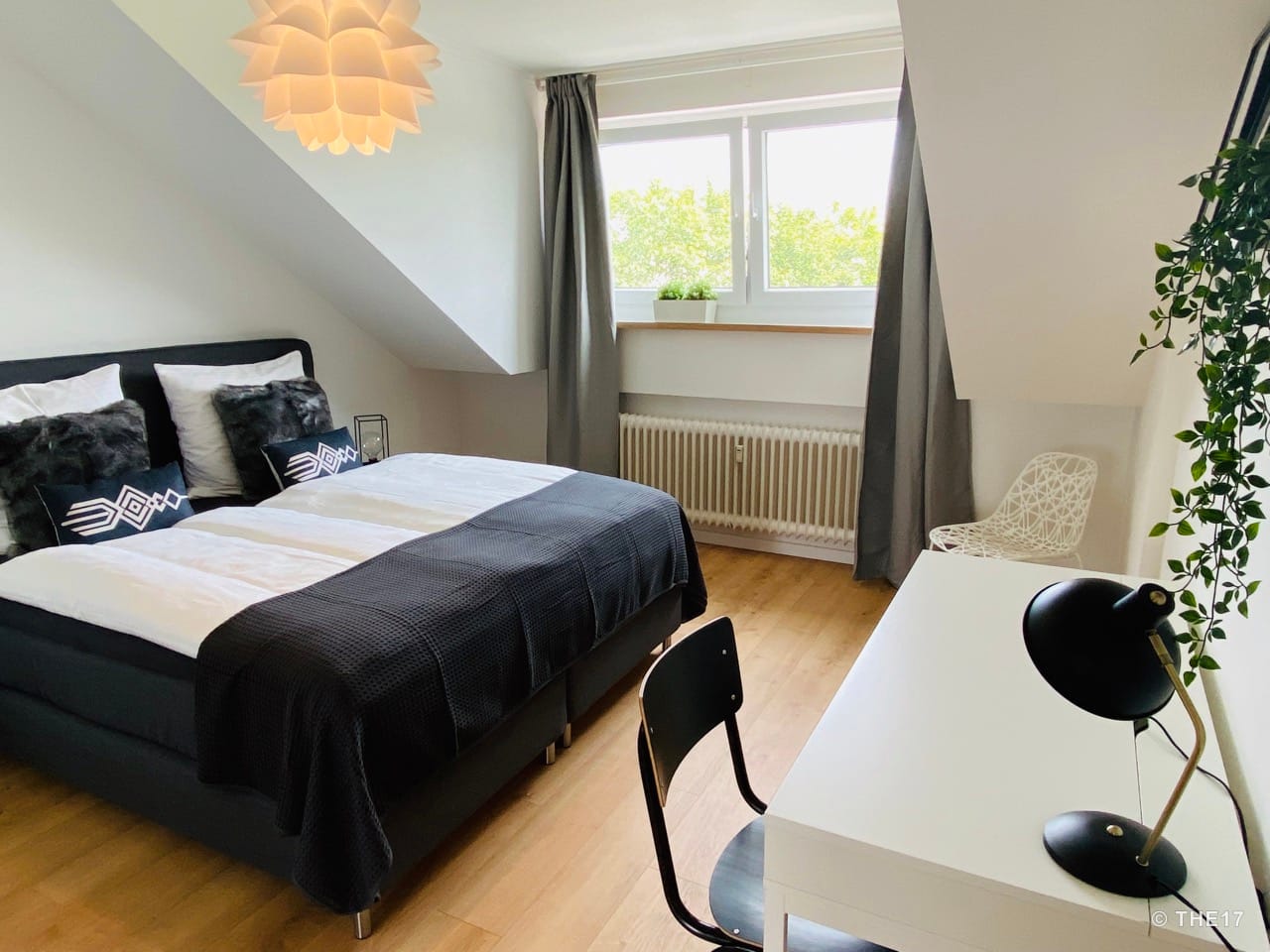 Bedroom with double bed, wooden floor, window with gray curtains, white desk, chairs, and hanging lamp.