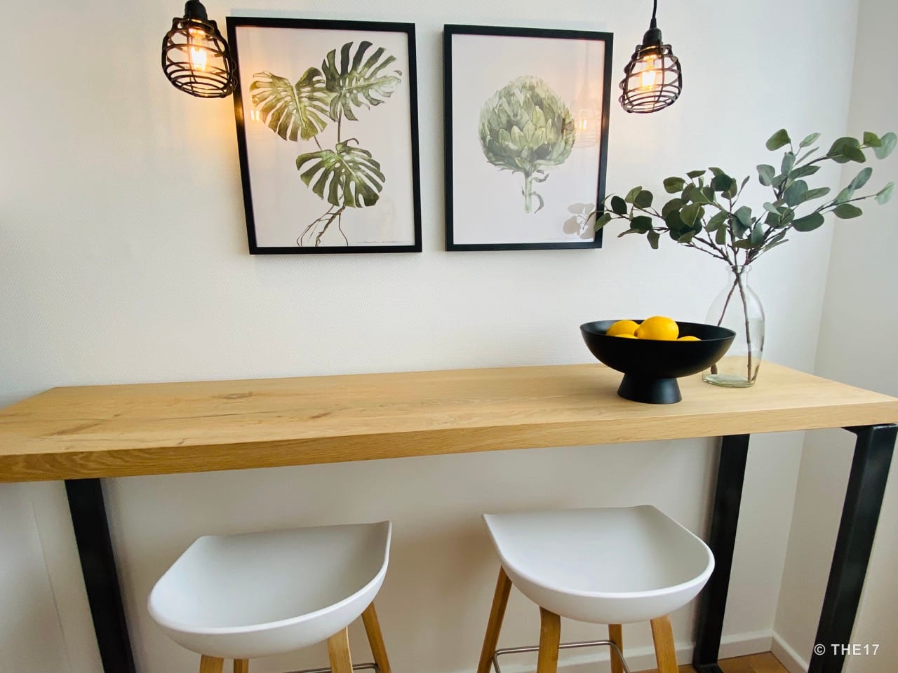 Modern breakfast bar with wooden top, two white chairs, two framed plant prints on the wall, and hanging lamps.