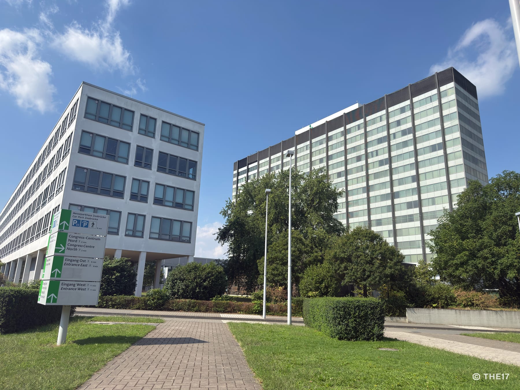 Zwei moderne Bürogebäude mit vielen Fenstern, umgeben von Bäumen und Grünflächen bei blauem Himmel.