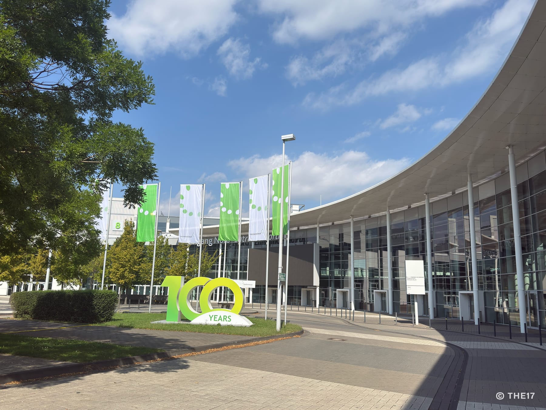 Eingang Nord der Koelnmesse in Köln-Deutz: geschwungene Glasfassade, davor Fahnen und die Skulptur „100 Years“.
