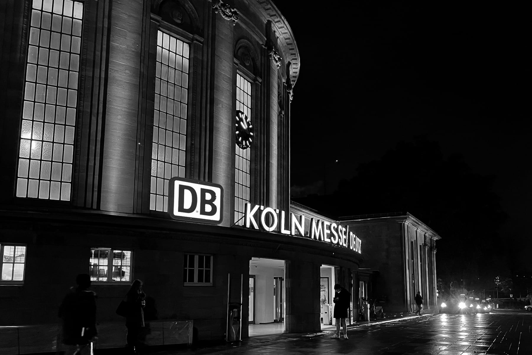 Nachtaufnahme des Eingangs eines Bahnhofs mit beleuchtetem DB-Logo und Schriftzug Köln Messe Deutz.