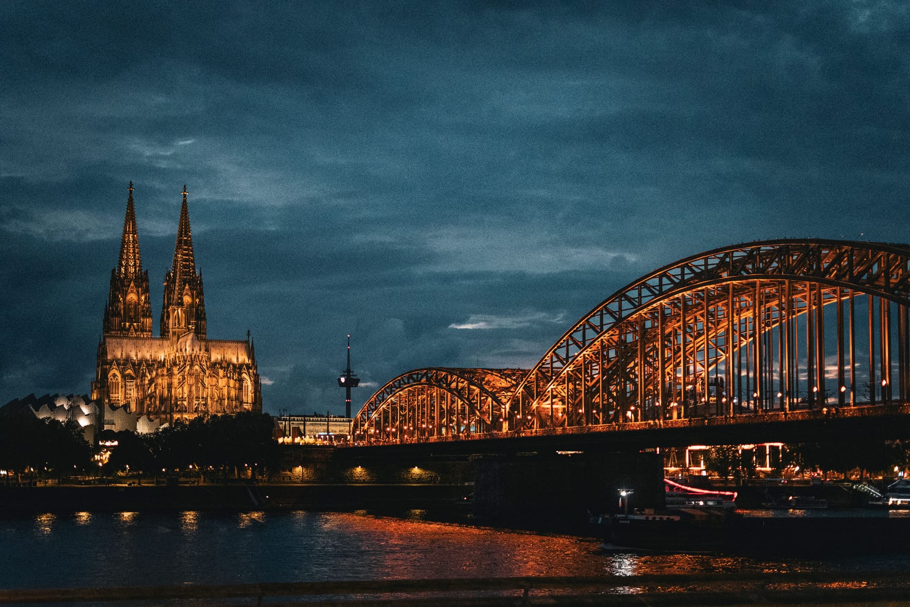 Beleuchteter Kölner Dom neben der erleuchteten Hohenzollernbrücke bei Nacht am Fluss.