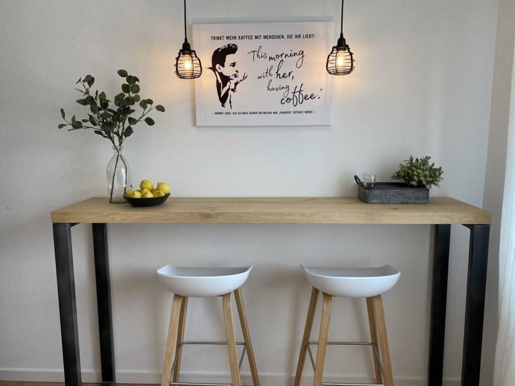 A minimalist dining area with a wooden bar table, on it a glass vase with green plant branches, a bowl with lemons and a metal tray with a small plant and two tea lights. Above the table hangs two industrial pendant lights. On the wall behind is a poster with a quote and illustration of Johnny Cash. In front of the table is two white bar stools with wooden legs.