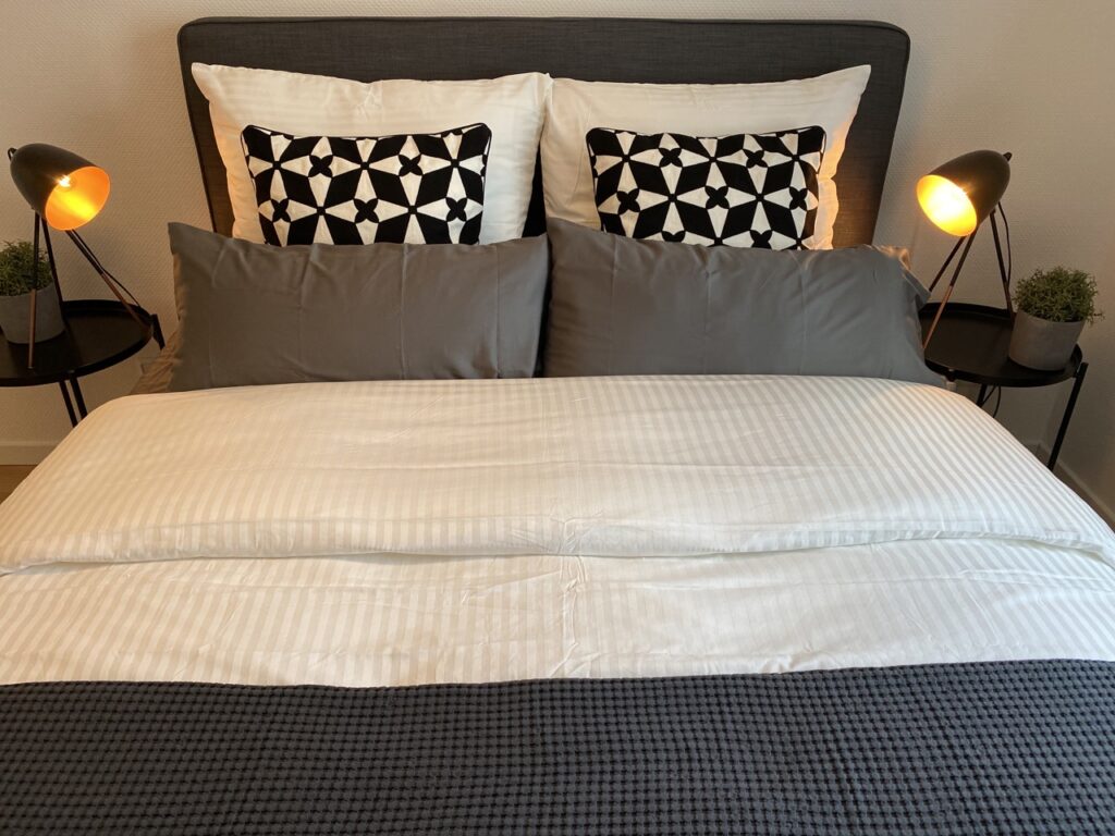 A neatly made bed with a dark gray headboard, white striped bedding and gray and black and white patterned pillows. On both sides of the bed are nightstands with modern black table lamps turned on and small green plants.