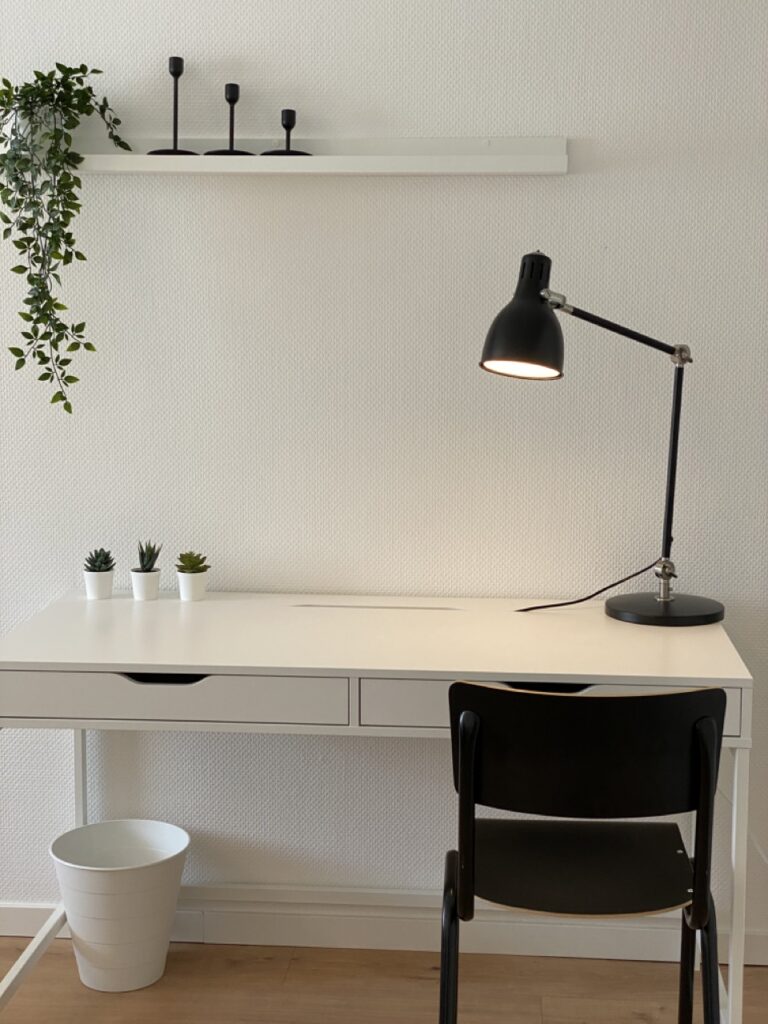 Workstation with a white desk on which stand three small succulents in white pots. Above the desk is a shelf with three black candlesticks. On the left side of the picture a climbing plant hangs down from the ceiling. Auf der rechten Seite steht eine schwarze Schreibtischlampe mit einem biegsamen Arm auf dem Schreibtisch. There is a black chair in front of the desk and a white wastebasket next to it. The room has a bright wallpaper and a wooden floor.