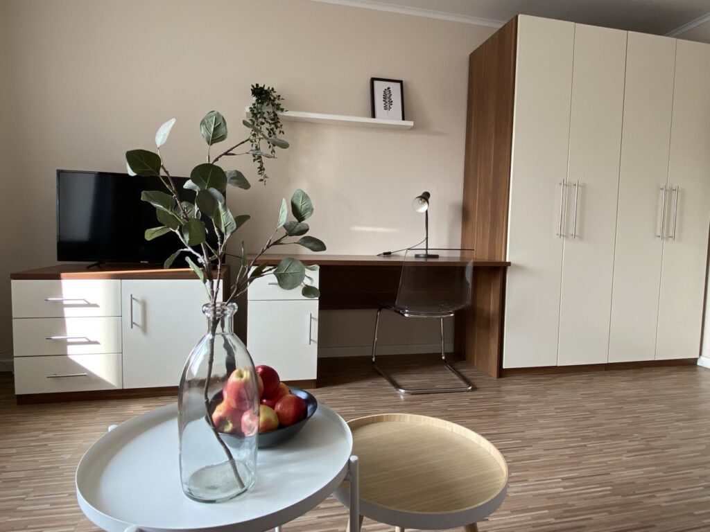 Modern furnished room with a flat screen TV on a white and brown sideboard. In front of the sideboard is a round white side table with a clear glass vase containing green plants and a black bowl with red apples. Next to the TV is a work area with a dark brown desk, a clear desk chair and a small desk lamp. On the right side of the picture is a large white closet with silver handles. The room has a wood-colored floor and light walls with a framed black and white picture on it.