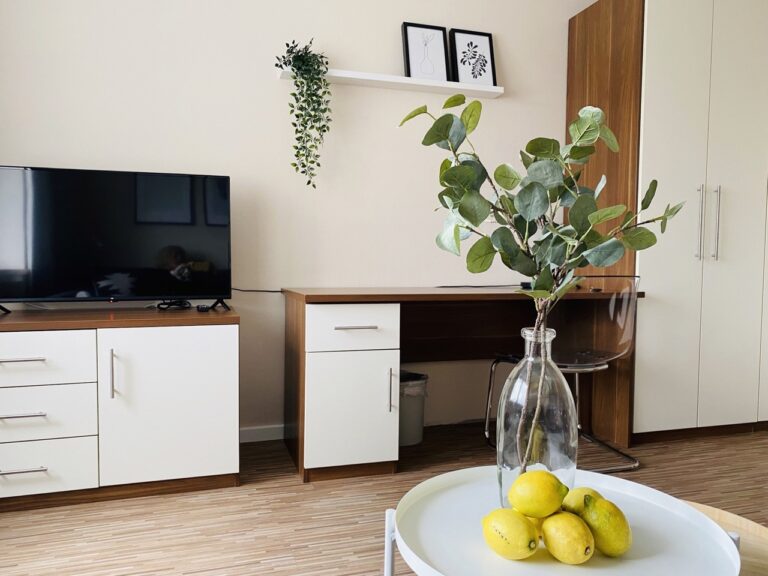 Modern furnished living room/bedroom with a large flat screen TV on a white piece of furniture with brown accents. Next to it is a matching dresser. A shelf with two black and white picture frames hangs on the wall. A green plant arrangement hangs vertically on the wall. In the foreground, a transparent vase with green leaves sits on a white table, with yellow lemons next to it. The floor is covered with light laminate.