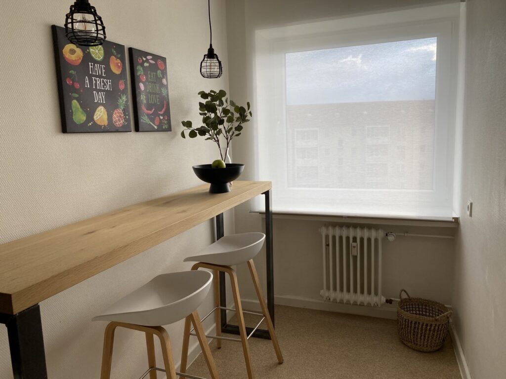 A cozy seating area in a kitchen with a high table made of wood and two modern bright bar stools under it. Above the table hang two black pendant lights and two framed pictures with fruit and vegetable motifs and the words 'Have a Fresh Day' and 'Eat Drink Love'. On the counter is a black bowl of apples and a green plant in a bottle. Next to the window is a radiator and below it is a woven basket on a light floor.