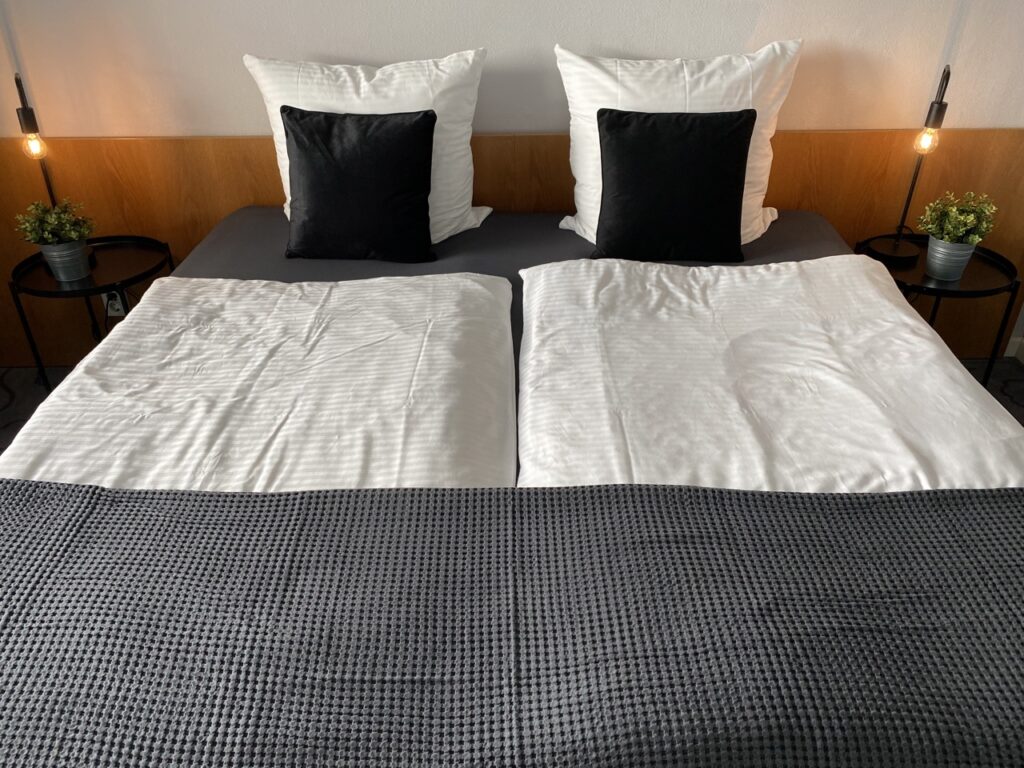 A neatly made double bed with white sheets and a dark gray comforter at the foot. There is a small black pillow on each pillow. On either side of the bed are bedside tables with black round tabletops, each with a lamp with an exposed light bulb and a small pot with green plants.