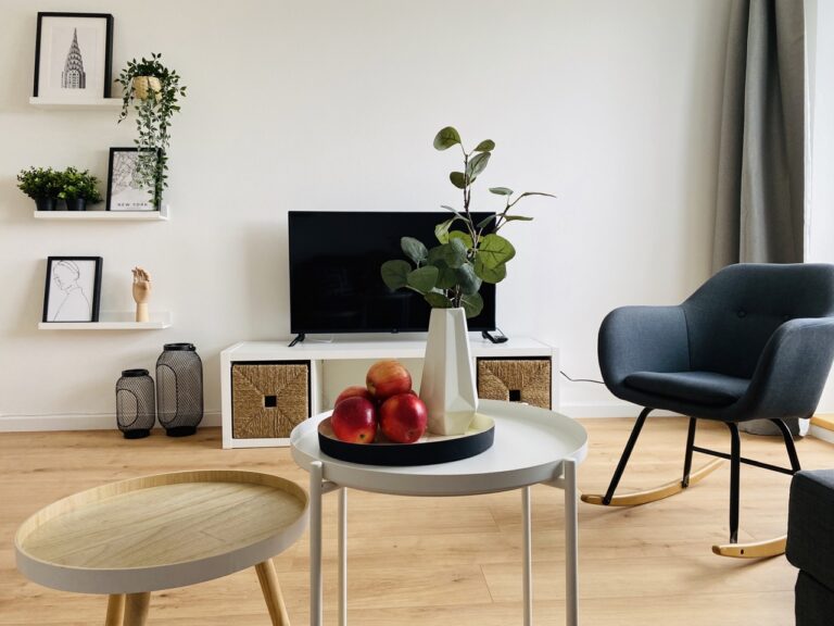 A modern furnished living room with a gray armchair, a white coffee table with apples, a TV on a low cabinet and decorative wall shelves displaying artwork and plants.